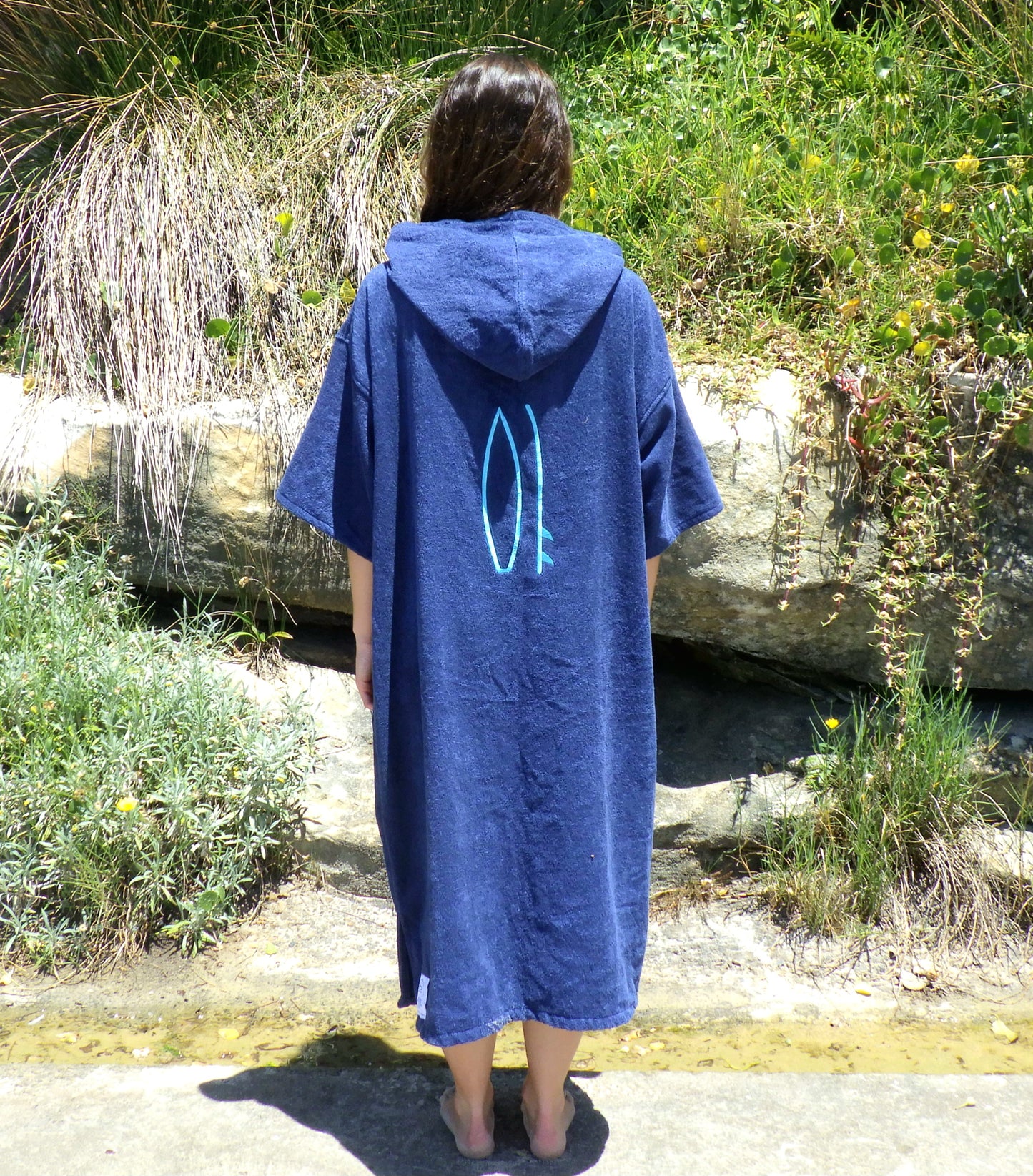 Person wearing a blue hooded towel with a design, standing outdoors near rocks and plants.
